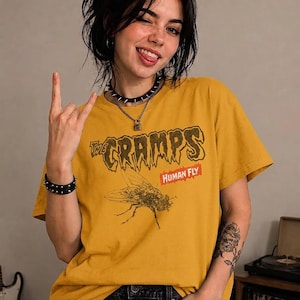 May include: A mustard yellow short-sleeved t-shirt featuring a graphic print of a housefly and the text "The Cramps" in a stylised font above the word "Human Fly" in a red rectangular box. The shirt has a crew neckline and a relaxed fit.