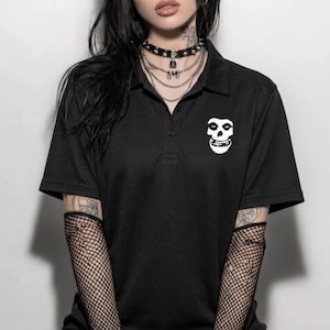 Może przedstawiać: Czarna koszulka polo z białym logo czaszki Misfits na piersi. Koszulka ma kołnierzyk i krótkie rękawy. Osoba nosi czarny naszyjnik typu choker i rękawy z siatki.
