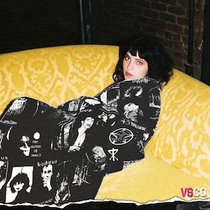 Op de afbeelding: Een persoon zit op een gele bank met een patroon en draagt een zwarte jas met witte afbeeldingen en tekst. De jas heeft afbeeldingen van muzikanten en bandlogo's. De achtergrond omvat een bakstenen muur en een gele bank.