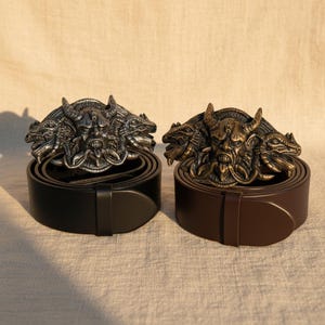Puede incluir: Dos cinturones de cuero, uno negro y otro marrón, con hebillas metálicas ornamentadas. Las hebillas presentan un diseño detallado de demonio de tres cabezas con cuernos y rasgos faciales intrincados. Los cinturones se exhiben sobre una superficie neutra.