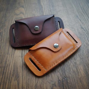 May include: Two leather pouches in shades of brown and tan rest on a wooden surface. Each pouch features a flap secured by a metal snap and has slots on the sides. The pouches are likely designed to hold small items.