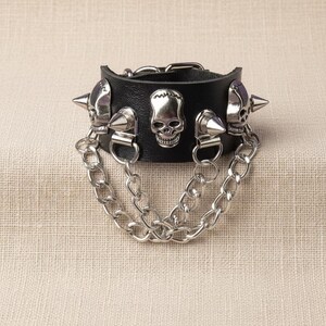 Puede incluir: Brazalete de cuero negro con detalles de calaveras y púas plateadas. El brazalete presenta tres diseños de calaveras y múltiples púas plateadas. Dos cadenas plateadas cuelgan del brazalete, lo que aumenta la estética punk rock.