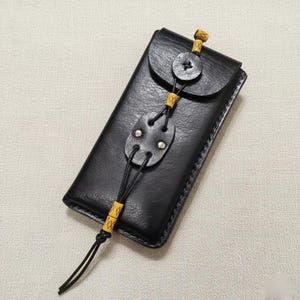 May include: Black leather phone case with a flap closure and button detail. The case features a decorative strap with yellow accents and a small, oval-shaped embellishment with silver studs. The stitching is visible around the edges.