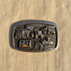 Puede incluir: Hebilla de cintur&oacute;n rectangular de metal con la palabra "CATERPILLAR" en la parte superior. La hebilla presenta un dise&ntilde;o en relieve de varios veh&iacute;culos de construcci&oacute;n, con el logotipo "CAT" en amarillo y negro.