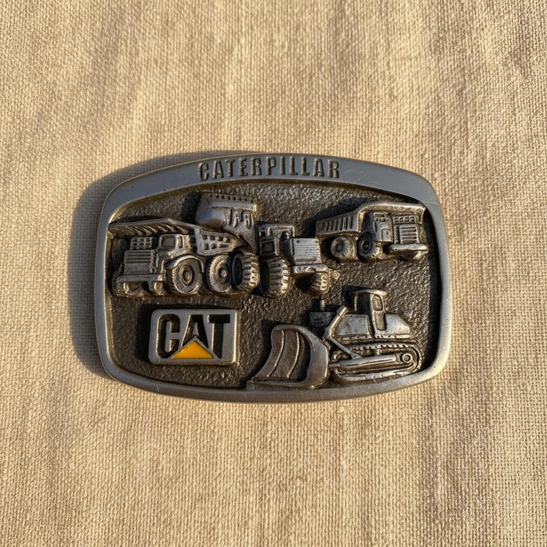 Puede incluir: Hebilla de cintur&oacute;n rectangular de metal con la palabra "CATERPILLAR" en la parte superior. La hebilla presenta un dise&ntilde;o en relieve de varios veh&iacute;culos de construcci&oacute;n, con el logotipo "CAT" en amarillo y negro.