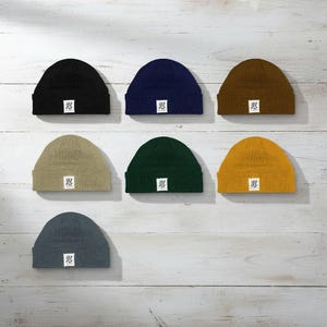 Puede incluir: Siete gorros en varios colores, incluyendo negro, azul marino, marrón, beige, verde, amarillo y gris. Cada gorro tiene un pequeño parche cuadrado blanco con un logotipo. Los gorros están sobre una superficie de madera clara.