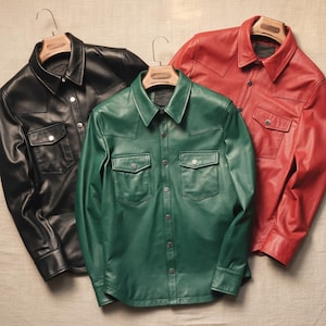 May include: Three leather shirts in black, green, and red hang on wooden hangers. Each shirt has two chest pockets and a button-down front. The shirts are displayed against a neutral background.