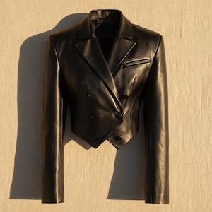 Puede incluir: Una blazer de cuero negro con diseño cruzado. La chaqueta presenta solapas con muescas, un pequeño bolsillo en el pecho y mangas largas. El largo recortado y el ajuste entallado crean una silueta moderna. La blazer tiene botones negros y un estilo clásico y atemporal.