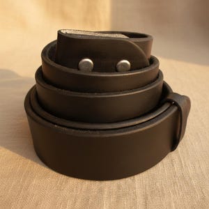 Puede incluir: Un cintur&oacute;n de cuero negro enrollado, con dos remaches plateados. El cintur&oacute;n est&aacute; hecho de cuero grueso y duradero. La hebilla no es visible en la imagen.