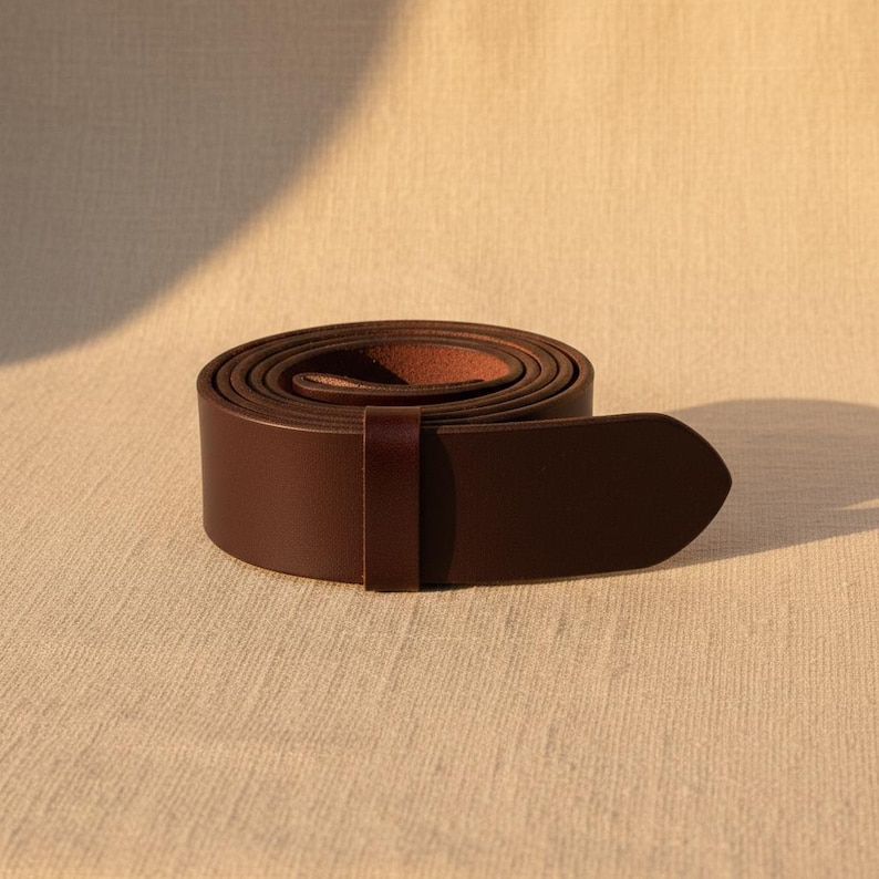 Puede incluir: Un cintur&oacute;n de cuero marr&oacute;n oscuro enrollado, con un extremo redondeado. El cintur&oacute;n es de color liso y parece estar hecho de material de alta calidad. La hebilla no es visible. El cintur&oacute;n est&aacute; sobre una superficie de tela beige.