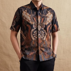 Puede incluir: Una camisa de manga corta con un intrincado estampado batik. La camisa presenta un fondo oscuro con un diseño detallado en tonos de marrón, naranja y crema. Tiene un cuello clásico y un bolsillo en el pecho.
