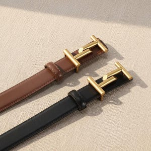 Pode incluir: Duas cintas de couro, uma castanha e outra preta, com fivelas de metal dourado. As fivelas têm um design distinto. As cintas são exibidas numa superfície de cor neutra.