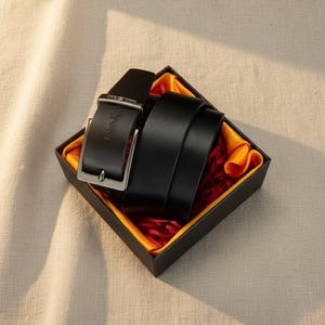 Op de afbeelding: Een zwarte leren riem met een zilveren gesp, gepresenteerd in een zwarte geschenkdoos met een gele en rode binnenkant. De riem is opgerold en klaar voor gebruik. De doos is vierkant en ligt op een neutraal gekleurd oppervlak.