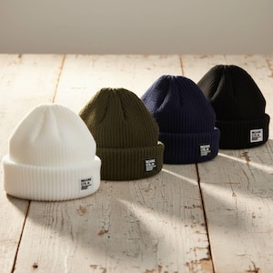 May include: Four ribbed knit beanies in white, olive green, navy blue, and black are displayed on a wooden surface. Each beanie has a folded brim and a small white tag with black text. The beanies are a casual accessory.