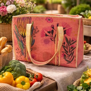 May include: A coral-colored tote bag with floral patterns and yellow handles sits on a wooden table. The bag is filled with groceries, including yellow bell peppers, tomatoes, and broccoli. Baskets of flowers and bread are in the background.