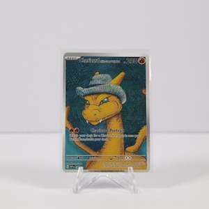 May include: A Charizard-Portrait trading card featuring a yellow dragon wearing a blue hat. The card has a dark blue background and is displayed on a clear plastic stand. The card's text includes "Charizard" and "-Portrait".