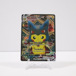 May include: A Pokémon trading card featuring Pikachu dressed as Lucario. The card has a gold, black, and blue design with the text "Lucario V". The card is displayed on a clear plastic stand.
