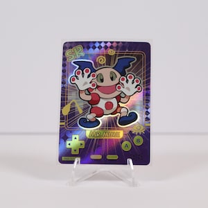 May include: A holographic trading card featuring Mr. Mime from the Pokémon series. The card has a purple background with a diamond pattern and a cartoon image of Mr. Mime. The card is displayed on a clear plastic stand.