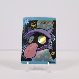 May include: A holographic trading card featuring the Pokémon Shellder. The card has a blue border and a black background with the Shellder character in the center. The character has a purple shell, black eyes, and a pink tongue.