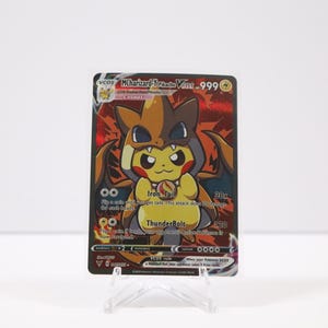May include: A Pokémon trading card featuring Pikachu dressed as Charizard. The card has a red and orange background with the text "M Charizard-Pikachu VMAX 999". The card is displayed on a clear plastic stand.