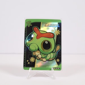 May include: A holographic trading card featuring the Pokémon Caterpie. The card has a green border with the name "CATERPIE" and a cartoon illustration of the Pokémon. The card is displayed on a clear plastic stand.