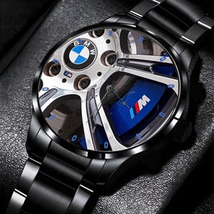 May include: Black wristwatch with a round face featuring a BMW wheel design. The watch face includes the BMW logo and the M logo. The watch has a black metal band and a black bezel.