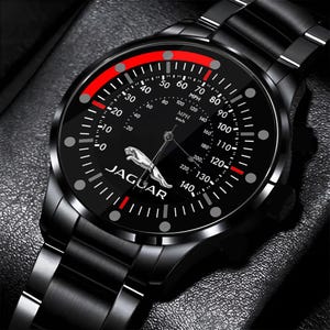 Pode incluir: Relógio de pulso preto com mostrador estilo velocímetro. O mostrador é preto com marcações brancas e vermelhas, incluindo indicadores de velocidade em MPH e km/h. O logotipo da Jaguar é exibido. A pulseira é de metal preto.