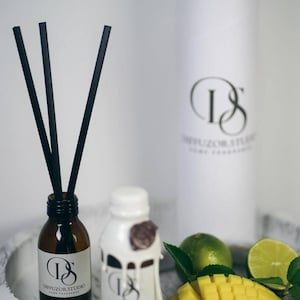 May include: Aromatherapy set with two small bottles, one amber and one white, labelled "Diffuzor Studio". Black reeds are in the amber bottle. A white cylinder and a wooden tray with mango and lime complete the scene.