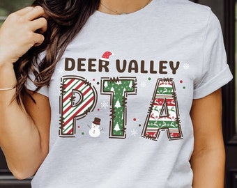 May include: White t-shirt with a Christmas-themed design that says "Deer Valley PTA". The letters are decorated with red and green stripes, snowflakes, and Christmas trees.