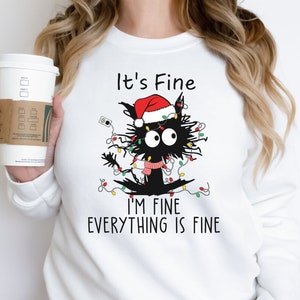 May include: White sweatshirt featuring a cartoon black cat tangled in Christmas lights and wearing a Santa hat and scarf. Text reads "It's Fine, I'm Fine, Everything is Fine."