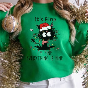 May include: A green Christmas sweater with a black cat wearing a Santa hat and tangled in Christmas lights. The text on the sweater reads 'It's Fine', 'I'm Fine', and 'Everything is Fine'.