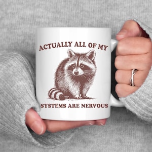May include: White ceramic mug with a brown illustration of a raccoon and the text "Actually all of my systems are nervous".
