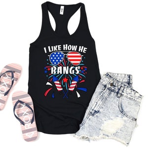 May include: Black tank top with the text "I like how he bangs" and a graphic of sunglasses in the American flag design. The tank top is paired with denim shorts and striped flip-flops.
