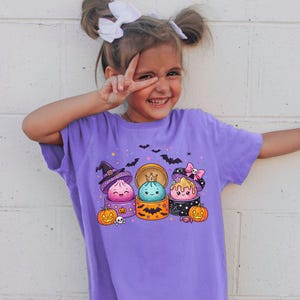May include: A violet short-sleeve t-shirt featuring a graphic print of three cartoon dumplings in bamboo steamers. The dumplings are decorated with a witch hat, a crown, and a pink bow, surrounded by orange pumpkins, bats, and small skulls.
