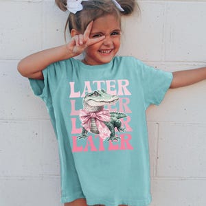May include: A turquoise t-shirt with the word "LATER" in pink, repeated three times, and a graphic of an alligator wearing a pink bow. The shirt is worn by a young person.