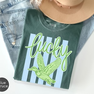 May include: A green t-shirt with the word "lucky" in a light green script, and a green duck graphic. The shirt is laid on top of blue jeans and a tan hat. A gold necklace is visible.