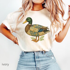 Puede incluir: Camiseta de color marfil con una ilustraci&oacute;n detallada de un pato. El pato tiene una cabeza verde, un pico amarillo y un cuerpo estampado en tonos azules, amarillos y marrones. El pato est&aacute; sobre hojas verdes. La palabra "ivory" est&aacute; impresa en la parte inferior.