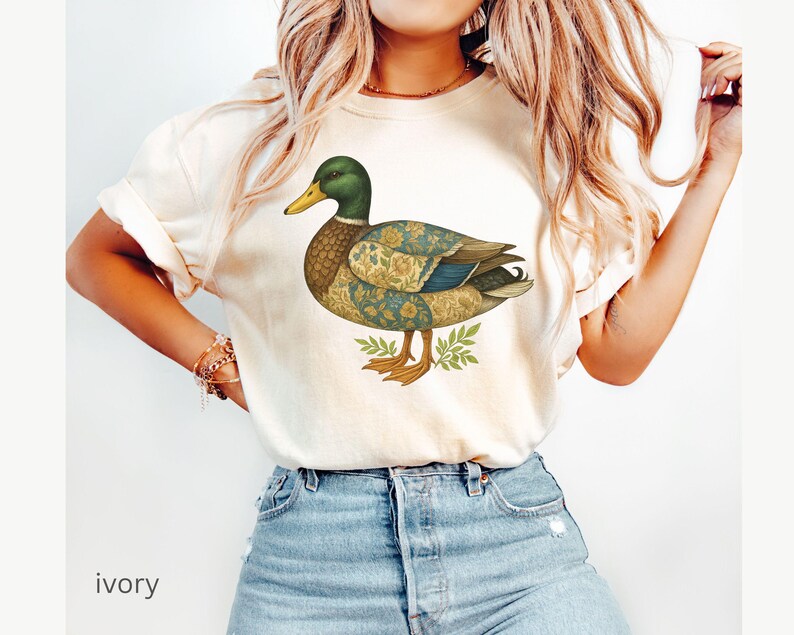 Puede incluir: Camiseta de color marfil con una ilustraci&oacute;n detallada de un pato. El pato tiene una cabeza verde, un pico amarillo y un cuerpo estampado en tonos azules, amarillos y marrones. El pato est&aacute; sobre hojas verdes. La palabra "ivory" est&aacute; impresa en la parte inferior.