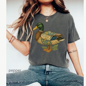 Puede incluir: Camiseta gris oscuro con una ilustraci&oacute;n detallada de un pato. El pato tiene una cabeza verde, pico amarillo y alas estampadas en azul, dorado y marr&oacute;n. La palabra "pepper" es visible en la parte inferior de la imagen.