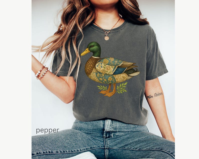 Puede incluir: Camiseta gris oscuro con una ilustraci&oacute;n detallada de un pato. El pato tiene una cabeza verde, pico amarillo y alas estampadas en azul, dorado y marr&oacute;n. La palabra "pepper" es visible en la parte inferior de la imagen.