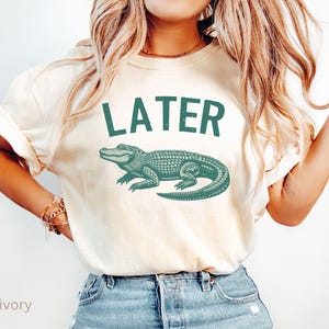 Op de afbeelding: Een wit T-shirt met de tekst "LATER" in zwart en een groene alligator-afbeelding. Het shirt wordt gedragen door een persoon met lang blond haar en is te zien vanaf de borst.