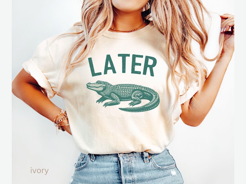 Op de afbeelding: Een wit T-shirt met de tekst "LATER" in zwart en een groene alligator-afbeelding. Het shirt wordt gedragen door een persoon met lang blond haar en is te zien vanaf de borst.
