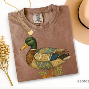 Puede incluir: Camiseta color espresso con una ilustraci&oacute;n detallada de un pato. El pato tiene una cabeza verde, un pico amarillo y alas estampadas con detalles florales y azules. Tambi&eacute;n se ven un collar con colgante de coraz&oacute;n dorado y un sombrero beige.