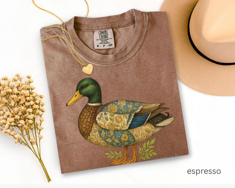 Puede incluir: Camiseta color espresso con una ilustraci&oacute;n detallada de un pato. El pato tiene una cabeza verde, un pico amarillo y alas estampadas con detalles florales y azules. Tambi&eacute;n se ven un collar con colgante de coraz&oacute;n dorado y un sombrero beige.