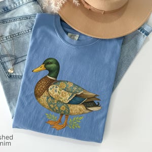 Puede incluir: Una camiseta azul claro con una ilustraci&oacute;n detallada de un pato, con cabeza verde, pico amarillo y alas estampadas. La camiseta est&aacute; colocada sobre unos vaqueros azules claros y cerca de un sombrero beige. El texto "washed denim" es visible.