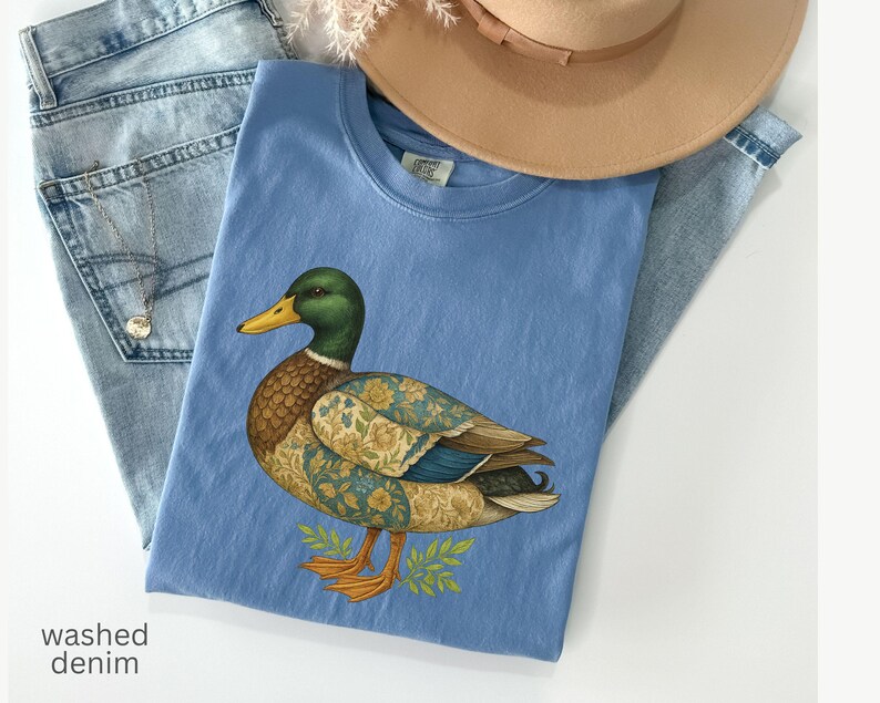 Puede incluir: Una camiseta azul claro con una ilustraci&oacute;n detallada de un pato, con cabeza verde, pico amarillo y alas estampadas. La camiseta est&aacute; colocada sobre unos vaqueros azules claros y cerca de un sombrero beige. El texto "washed denim" es visible.