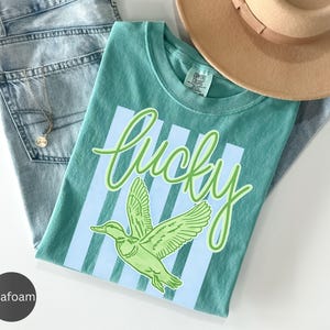 May include: Seafoam green t-shirt with the word "Lucky" in cursive and a green duck graphic. The shirt is laid on top of light blue jeans and a tan hat. A gold necklace is visible.