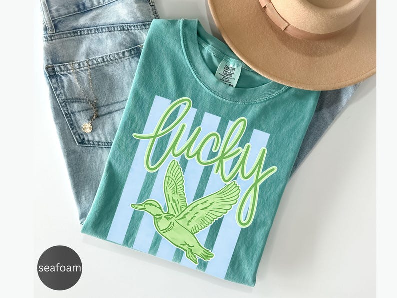 May include: Seafoam green t-shirt with the word "Lucky" in cursive and a green duck graphic. The shirt is laid on top of light blue jeans and a tan hat. A gold necklace is visible.