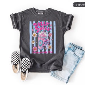 May include: A dark gray t-shirt with a graphic of a pink drink, daisies, and the text "Dilly Dolly". The shirt is paired with light blue denim shorts and black and white checkered slip-on shoes.