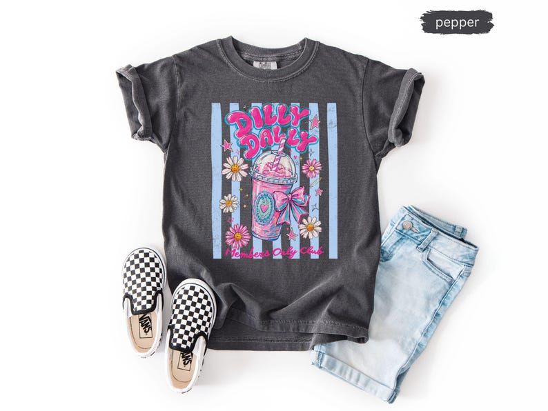 May include: A dark gray t-shirt with a graphic of a pink drink, daisies, and the text "Dilly Dolly". The shirt is paired with light blue denim shorts and black and white checkered slip-on shoes.
